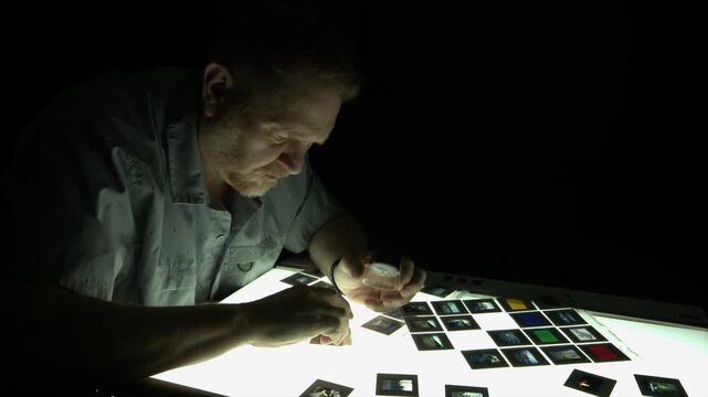 Male editor illuminated from lightbox in dark room, looks at slides
