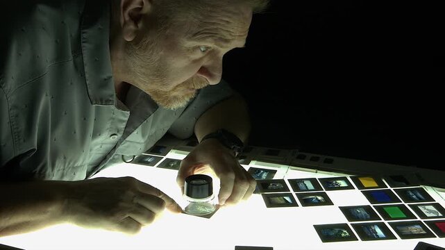 Creative editor lit by lightbox from below, inspects slide film in dark