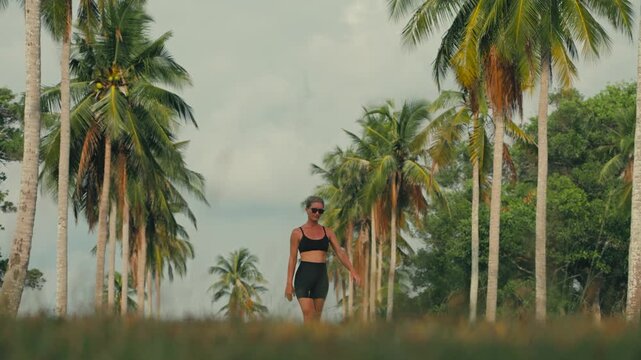 Fit Caucasian blonde woman walking happily and nonchalantly along a palm tree path in tropical Thailand. Relaxed travel lifestyle scene with lush greenery and warm exotic atmosphere