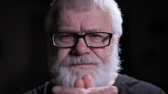 An elderly man in glasses, with gray hair and a beard, is immersed in prayer