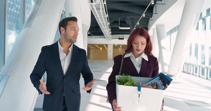 Male executive gestures supportively to sad dismissed woman carrying personal belongings box walking through bright modern office corridor