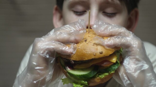 A boy in protective gloves looks hungrily at a large hamburger. Close-up of a child preparing to eat a juicy burger