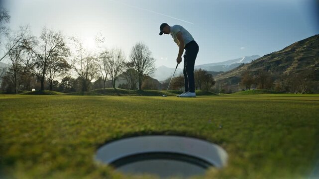 Wide shot of a golfer attempting a putt on the green in Sion, Switzerland, narrowly missing the hole. Calm golf course setting, scenic alpine surroundings, and authentic sports moment.