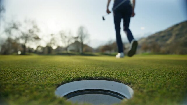 Golfer picking the ball from the hole on the green in Sion, Switzerland, then walking away after the putt. Calm golf course atmosphere, scenic alpine surroundings, and authentic sports moment.