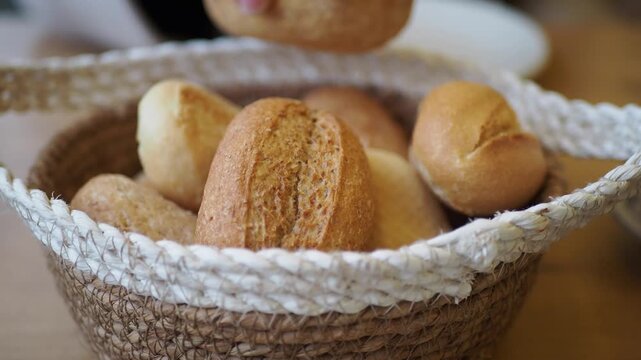 A close-up shot of a woven basket filled with freshly baked bread rolls, a person's hand reaches in and picks up a roll