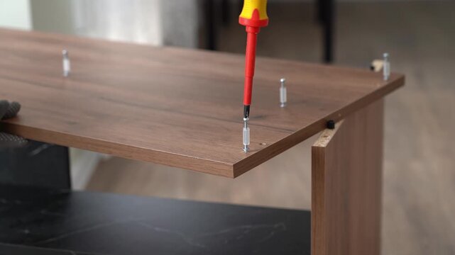 Male hands assemble nightstand using screwdriver. Tightening connecting bolt. Process of assembling furniture. Furniture assembler installs fasteners