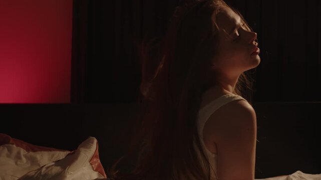 A beautiful tattooed woman wearing white lingerie sits on her hands and knees in bed, she throws her head backwards and waves her long hair in slow motion