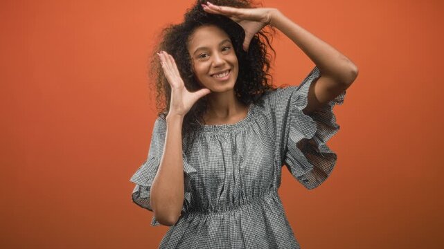 Teenage woman with curly hair in ruffled gingham dress frames her face with hands while smiling at camera in studio; youthful joy.