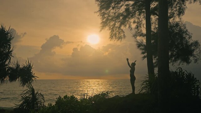 Silhouette of a woman transitioning from upward salute yoga pose to prayer hands under a tree near the sea during a beautiful golden sunset. Peaceful wellness moment by the coast