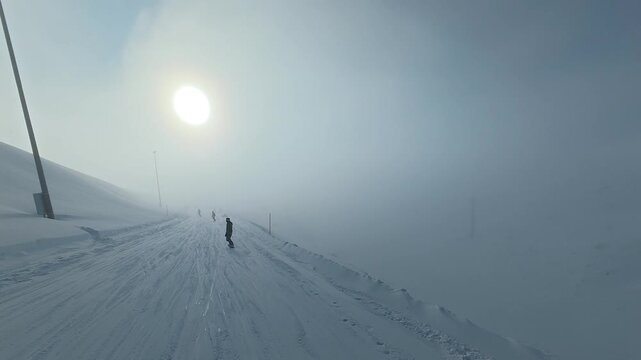 POV snowboarding down a slope through fog under bright sun and clouds. Action camera footage capturing speed, changing visibility, and atmospheric winter mountain conditions.