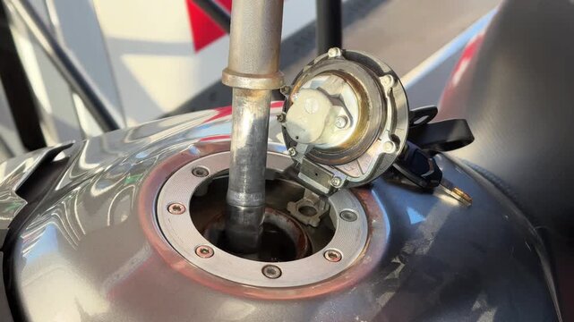 A man refuels his motorcycle, pouring gasoline through the tank opening in close-up. The scene captures the precise moment of filling up the gas tank.