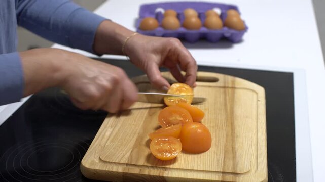 A man uses a knife to cut tomatoes on a wooden board while eggs lie in a box nearby on the kitchen counter.