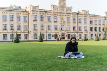 Obraz premium A student sits alone on a wide green lawn in front of a classic university building using a tablet. Campus landscape, solitary study, modern education