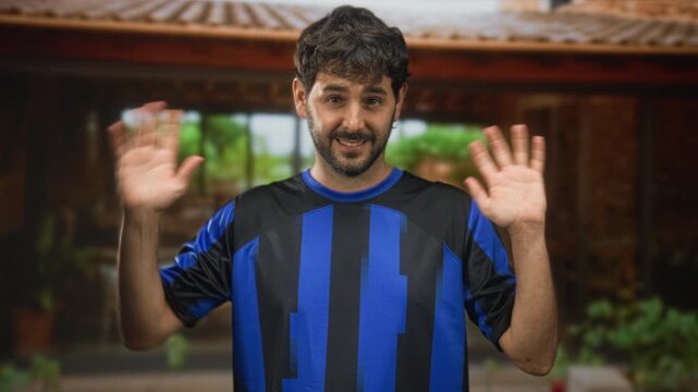 Man with beard waves right hand in front of a building wearing blue and black football jersey and earring, smiling into camera; friendly greeting.