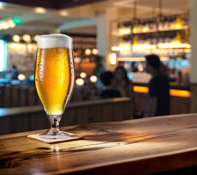 Glass of chilled beer on wooden bar tabletop and blurred brightly lit bar interior in the background.