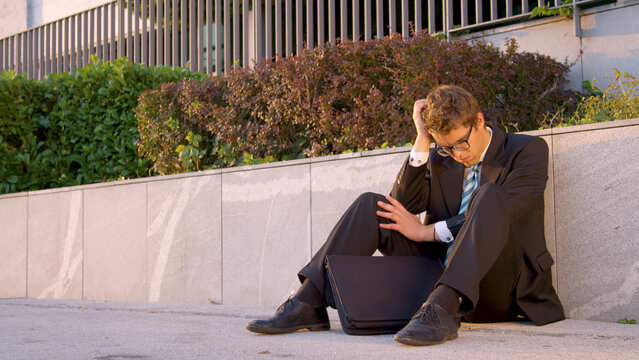 Depressed young Caucasian yuppie sitting on the sidewalk after going bankrupt.
