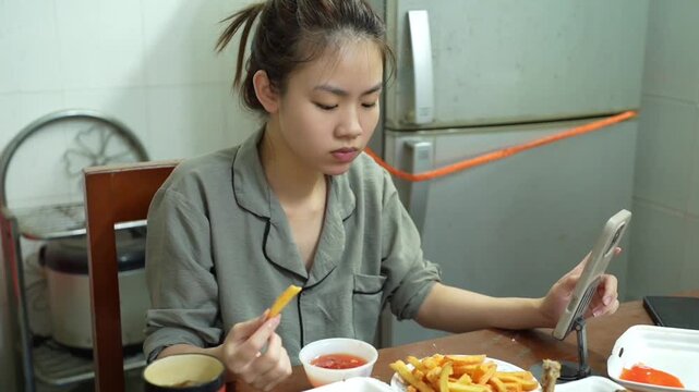 Young Asian woman eating fried food while using smartphone at home