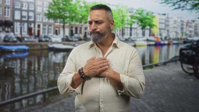 Man with hands clasped to chest on a street by an amsterdam canal wearing a beige linen shirt and beaded bracelets; quiet reflection.