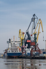 Obraz premium Cargo ship is docked at a busy port, featuring large cranes and industrial structures along the waterfront with a clear blue sky above