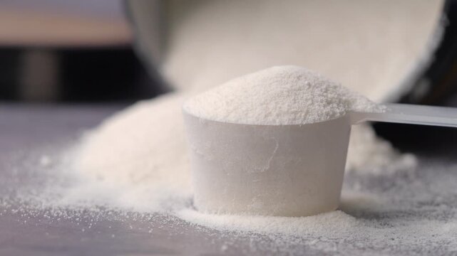 Slow motion close up of white protein powder in scoop on grey background with container blurred behind