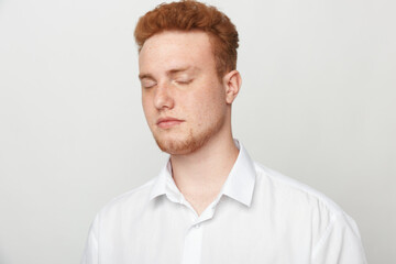 Fototapeta premium Young man with red hair and beard wearing white shirt closes eyes in calm expression. Isolated on white background. Concept of relaxation, peace, and mindfulness in portrait photography.