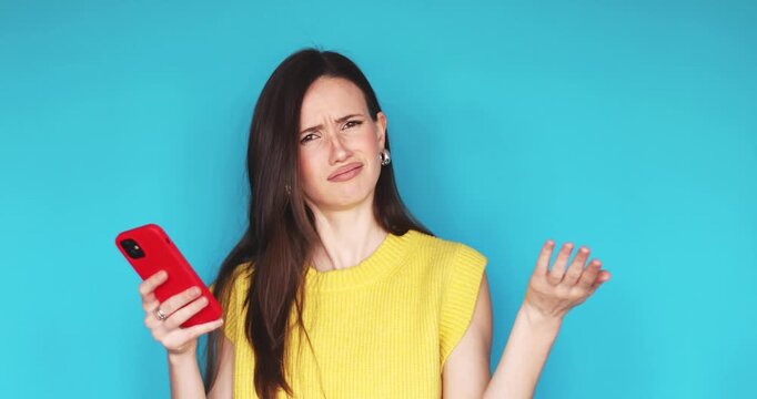 Surprised young woman looking at smartphone screen with disbelief and shock, raising hands in confusion, wearing yellow sweater against blue studio background