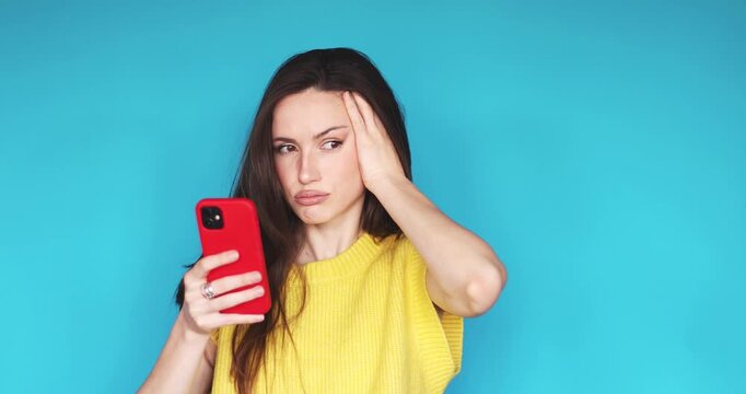 Frustrated young woman holding smartphone and looking at screen with headache and disappointment, massaging temple due to bad news or phone error, standing against blue studio background