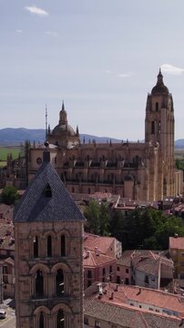 Aerial view across historic rooftops toward Iglesia de San Esteban and Segovia Cathedral with distant mountain landscape. Vertical