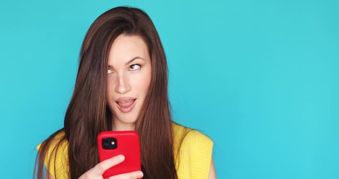 Brunette woman with long hair looking at smartphone screen typing message and making grimace of disgust isolated blue background negative reaction internet trolling social media content video portrait