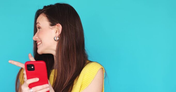 Cheerful young brunette woman looking at smartphone screen laughing and pointing finger at copy space while posing on blue background influencer sharing amazing mobile app or social media content