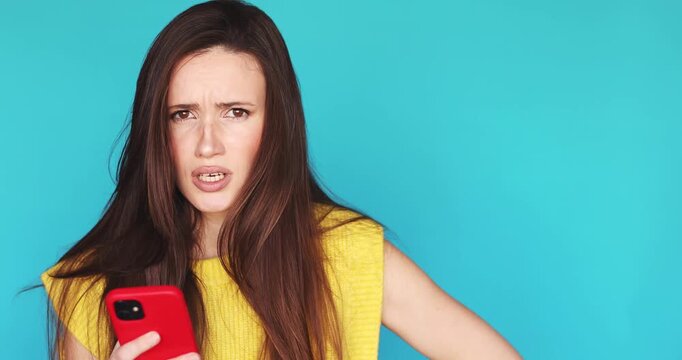 Confused and skeptical young woman looking at her red smartphone with disbelief and doubt while standing isolated on a vibrant cyan studio background expressing negative emotions and mistrust