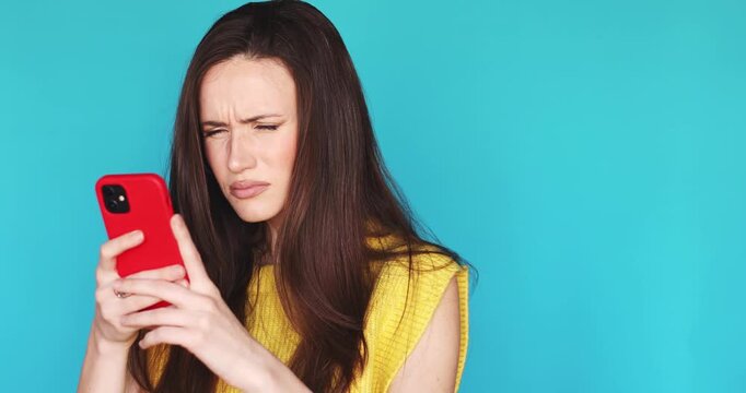 Annoyed and confused young woman looking at her smartphone with a skeptical facial expression and squinting eyes while standing against a blue background feeling irritated by a message