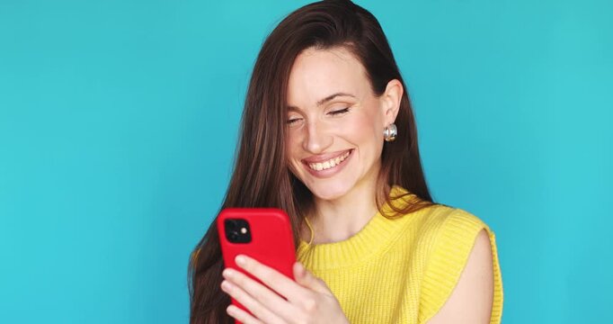 Happy young woman with long brown hair smiling and laughing while using red smartphone in hands, typing message or scrolling social media, posing in yellow sweater on vibrant blue studio background.