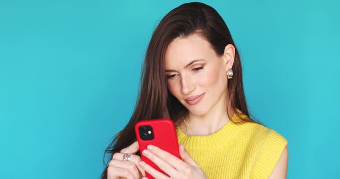 Happy young woman scrolling social media feed on red smartphone and smiling. Cheerful female user browsing internet or typing message in yellow sweater, posing on vibrant blue studio background.