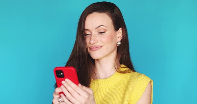 Confident young woman in yellow sweater looking at smartphone and nodding head in agreement. Cheerful female user saying yes with head gesture while holding red phone on blue studio background.