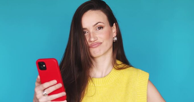 Skeptical young woman in yellow sweater looking at smartphone and shaking head in disagreement. Disappointed female user saying no with head gesture while holding red phone on blue background.