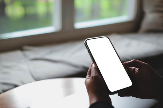 A close-up view of a person holding a smartphone with a blank white screen over a wooden table near a bright window in a cozy indoor setting.
