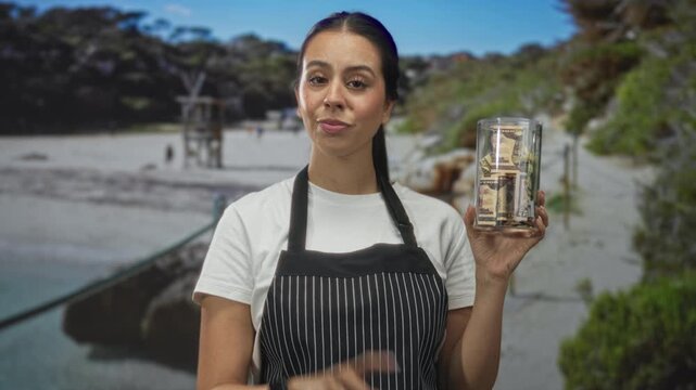 Young brunette woman in striped apron holding glass tip jar of cash at beach while pointing index finger to sky; charity generosity.