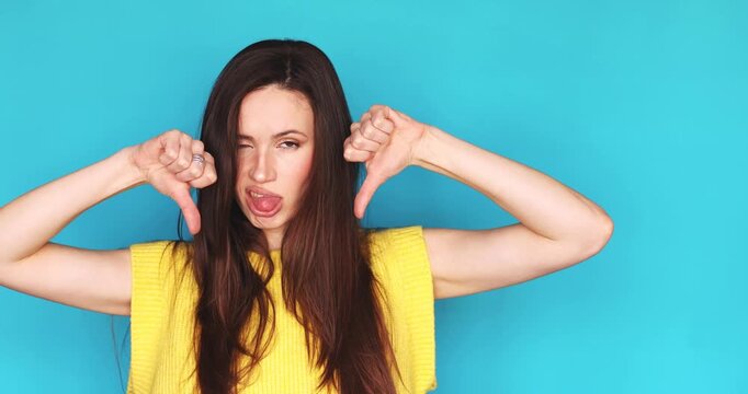 Dissatisfied young woman showing double thumbs down gesture and sticking out tongue in disgust isolated on blue background, unhappy female brunette expressing strong dislike and rejection