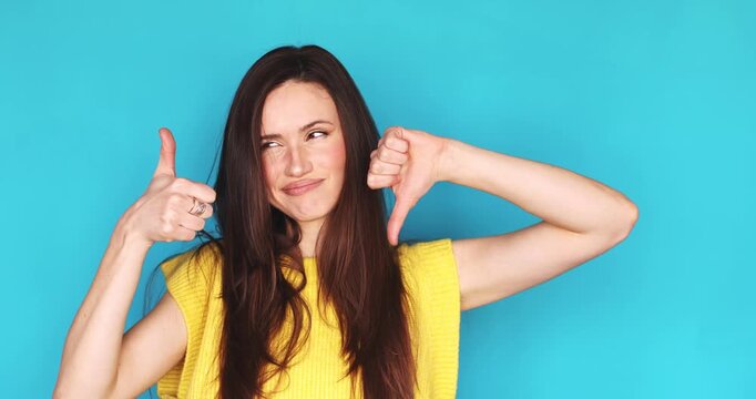 Expressive young woman showing thumb up and thumb down at the same time looking at camera with hesitant face isolated on blue background, female brunette comparing pros and cons, good or bad choice