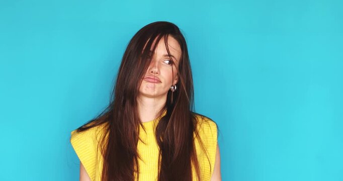 Bored and tired young brunette woman looking at camera with indifferent and annoyed face expression isolated on blue background, unhappy female feeling exhausted or unmotivated, close up portrait