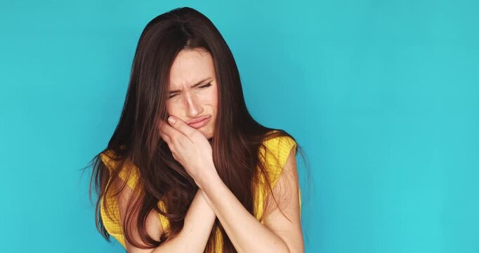 Young woman suffering from severe toothache and touching her cheek in pain isolated on blue background, unhappy female brunette feeling dental problem or sensitive teeth, medical concept