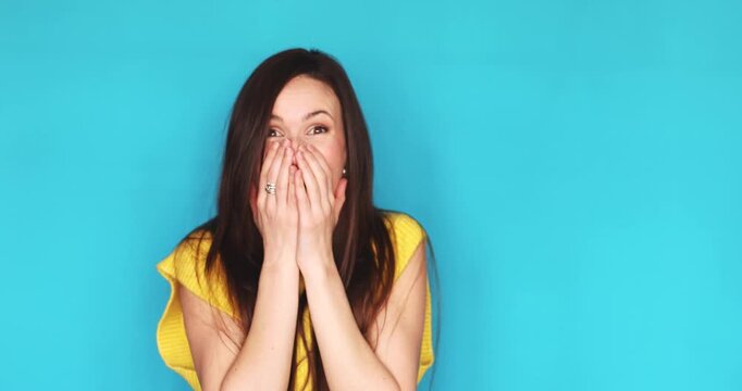 Excited and shocked young woman laughing and covering mouth with hands in disbelief isolated on blue background, happy female brunette celebrating success or great news, vivid emotions