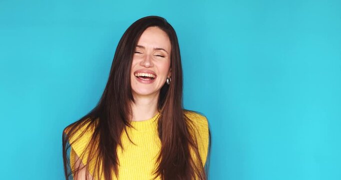 Excited young brunette woman in yellow knitted sleeveless top on turquoise background happily pointing directly at camera, laughing and beckoning to join, expressing selection, invitation and calling.