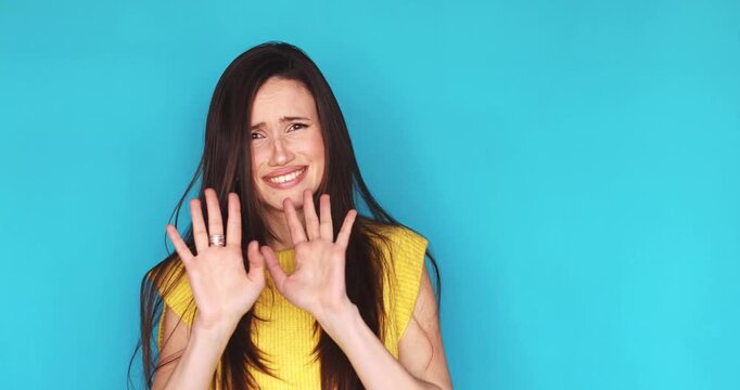 Young brunette woman in yellow top on turquoise background expressing disgust and strong refusal with facial expressions, saying no, strictly rejecting and strictly avoiding with hand gestures.