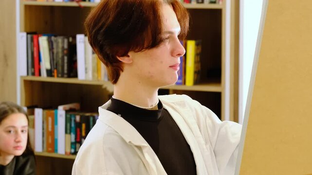 Young man student talks looking at whiteboard in school library. Teenage scholar explains lesson topic to classmates in college book storage