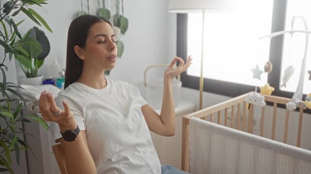 Woman sits with hands in meditation mudra and eyes closed beside a wooden baby cradle in a building nursery; mindfulness calm.