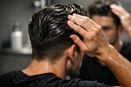 Man applying hair gel to wet dark hair in front of a mirror, close-up shot