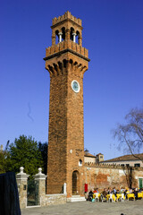 Fototapeta premium The Clock Tower in Murano, Venice, Italy, with people dining outdoors under a clear blue sky.