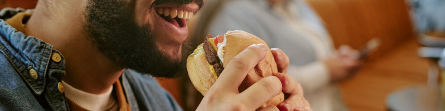 Header of young adult man smiling while holding hamburger near mouth, preparing to take bite, sitting indoors with blurred middle aged woman using smartphone in background